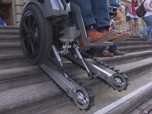 Ein Rollstuhl mit darunter gebauten Raupen fährt rückwärts eine Treppe hinauf. Ein Rollstuhl mit darunter gebauten Raupen fährt rückwärts eine Treppe hinauf.