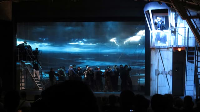 Auf der Leinwand im Hintergrund ein Gewitter auf dem Meer, davor Darsteller Auf der Leinwand im Hintergrund ein Gewitter auf dem Meer, davor Darsteller