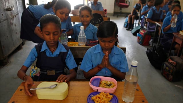 Indisches Klassenzimmer mit zwei Schülerinnen im Vordergrund; eine betet vor dem Essen. Indisches Klassenzimmer mit zwei Schülerinnen im Vordergrund; eine betet vor dem Essen.
