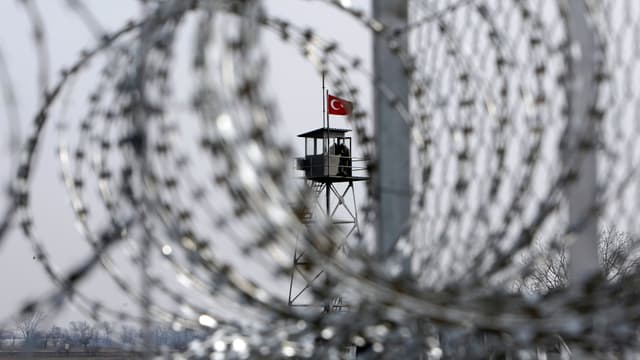 Ein Wachturm mit türkischer Flagge ist hinter einem Zaun mit Stacheldrahtrollen zu sehen.