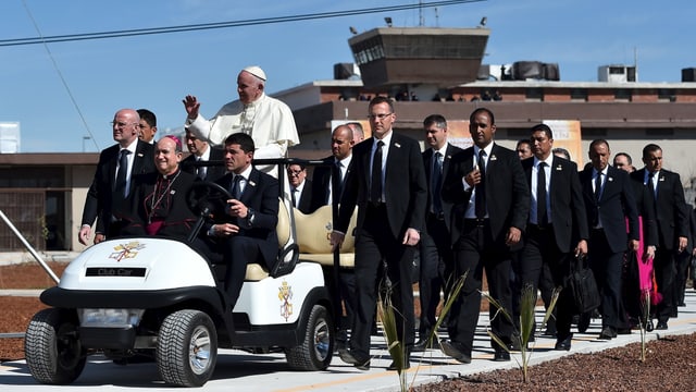 Papst stehend auf einem kleinen Auto, dahinter Männer in schwarzen Anzügen. Papst stehend auf einem kleinen Auto, dahinter Männer in schwarzen Anzügen.