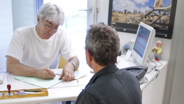 Arzt schreibt, Patient sitzt gegenüber. Arzt schreibt, Patient sitzt gegenüber.