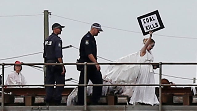 Eine Frau in einem weissen Kleid hält ein Schild «coal kills», sie wird von zwei Polizisten gefolgt. Eine Frau in einem weissen Kleid hält ein Schild «coal kills», sie wird von zwei Polizisten gefolgt.