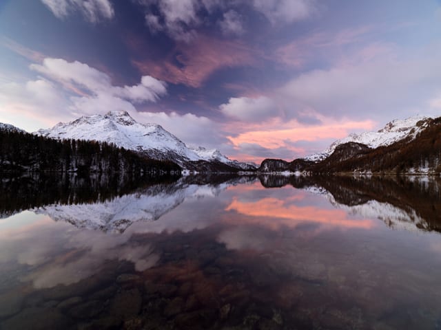 Im Vordergrund ein spiegelglatter Bergsee, in dem sich die verschneiten Talflanken und der Morgenhimmel spieglen.