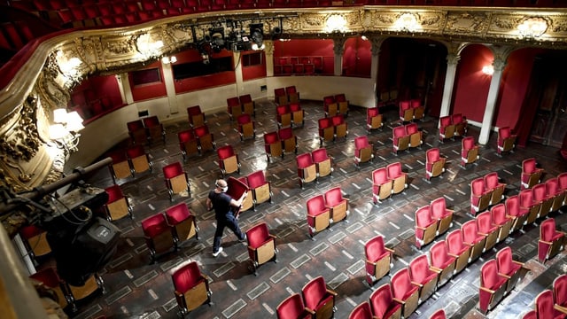 Ein Mann trägt einen roten Plüschsitz aus einem grossen Zuschauerraum, in dem die Sitzreihen durch Lücken unterbrochen werden. Ein Mann trägt einen roten Plüschsitz aus einem grossen Zuschauerraum, in dem die Sitzreihen durch Lücken unterbrochen werden.