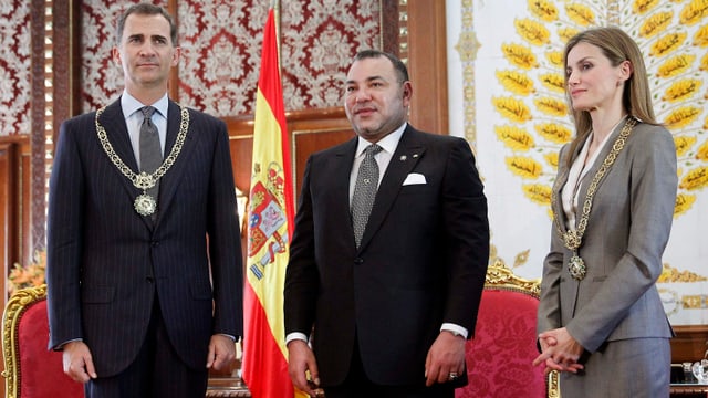 Felipe, Mohammed und Letizia stehen stramm vor roten Sesseln und einer spanischen Flagge. Felipe, Mohammed und Letizia stehen stramm vor roten Sesseln und einer spanischen Flagge.