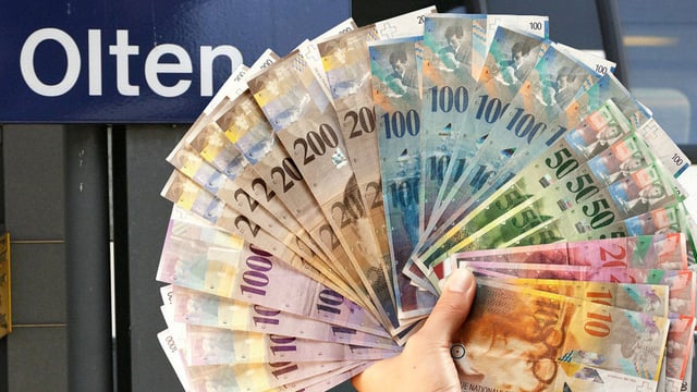 Hand hält Bündel von Geldscheinen vor Bahnhofsschild Hand hält Bündel von Geldscheinen vor Bahnhofsschild