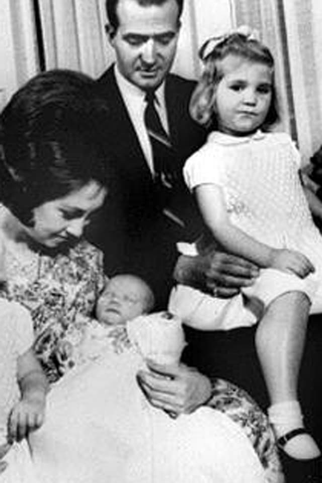 Familienfoto in schwarzweiss: Königin Sophia hält Baby Felipe auf dem Arm. Im Hintergrund sitzt seine Schwester in weissem Kleid auf der Schoss von König Juan Carlos.