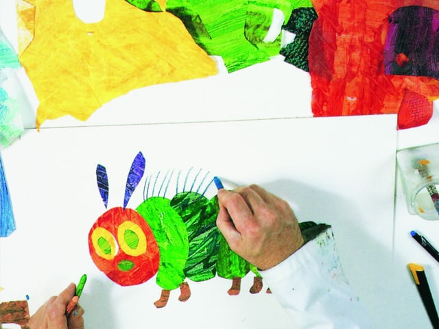 Hände auf einem Schreibtisch, auf dem farbiges ausgeschnittenes Papier und eine Skizze einer Raupe liegen. Hände auf einem Schreibtisch, auf dem farbiges ausgeschnittenes Papier und eine Skizze einer Raupe liegen.