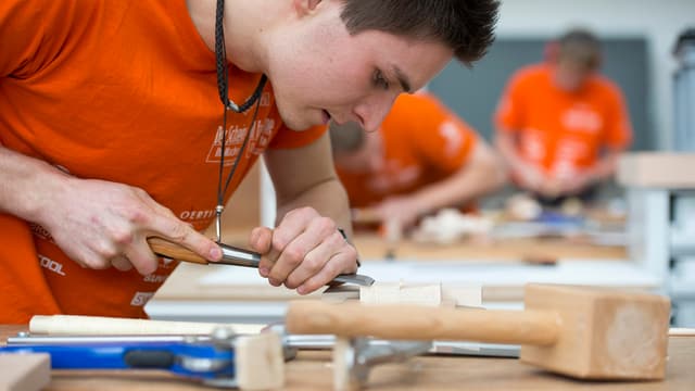 Lehrling bearbeitet an seinem Arbeitsplatz mit einem Werkzeug ein Stück Holz. Lehrling bearbeitet an seinem Arbeitsplatz mit einem Werkzeug ein Stück Holz.