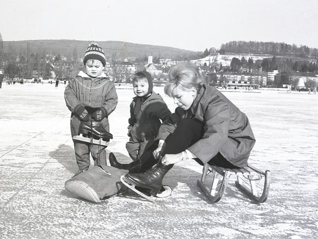 Ein Schwarz-Weiss Bild aus 1964 mit einer Frau und zwei Kinder auf einem gefrorenem See.