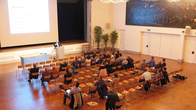 In einem Saal doziert ein Mann vor einer beleuchteten Leinwand, einige Zuhörer sitzen auf Stühlen. Viele Sitze bleiben aber leer. In einem Saal doziert ein Mann vor einer beleuchteten Leinwand, einige Zuhörer sitzen auf Stühlen. Viele Sitze bleiben aber leer.