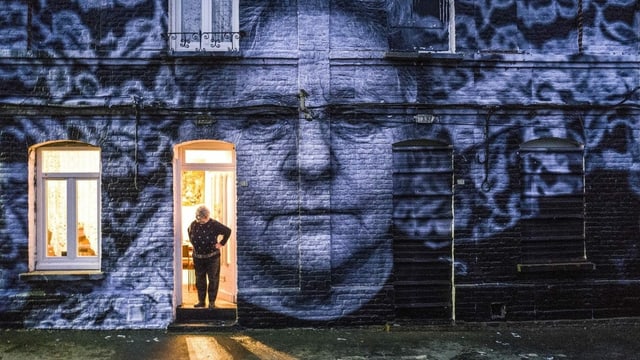 Ein Porträt einer Frau klebt an einer Fassade. Die Frau selbst steht in der Tür. Ein Porträt einer Frau klebt an einer Fassade. Die Frau selbst steht in der Tür.