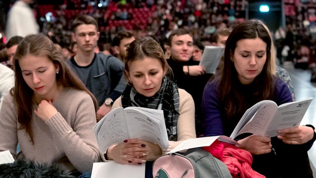 Drei junge Frauen sitzen in einem grossen Raum mit anderen, sie halten Papiere in der Hand. Drei junge Frauen sitzen in einem grossen Raum mit anderen, sie halten Papiere in der Hand.