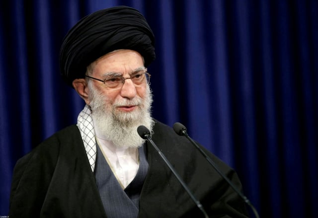 Ayatollah Khamenei bei einer Konferenz. Er trägt einen weissen Bart, eine Brille und religiöse Kopf- und Körperbedeckungen.