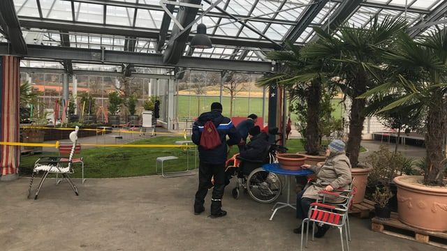Ein Glashaus, darin laufen Menschen herum, eine Frau sitzt an einem roten Tischchen. Ein Glashaus, darin laufen Menschen herum, eine Frau sitzt an einem roten Tischchen.