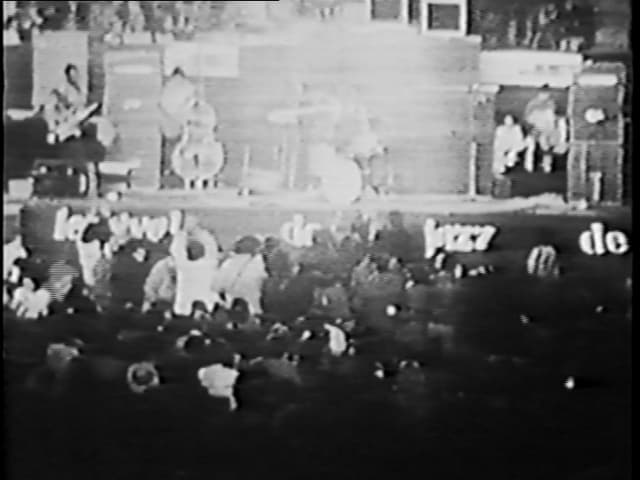 Schwarz-weisses Filmstill, Konzertbühne, Band im Hintergrund, Publikum im Vordergrund Schwarz-weisses Filmstill, Konzertbühne, Band im Hintergrund, Publikum im Vordergrund