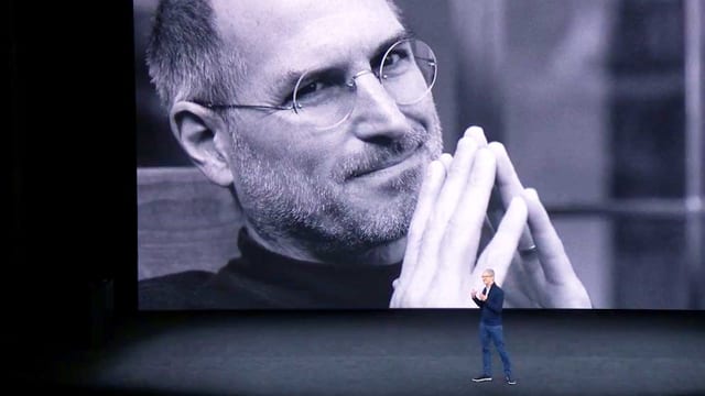Eine Nahaufnahme von Steve Jobs wird auf eine grosse Wand projiziert. Davor steht ein Mann in Jeans und hält eine Präsentation. Eine Nahaufnahme von Steve Jobs wird auf eine grosse Wand projiziert. Davor steht ein Mann in Jeans und hält eine Präsentation.