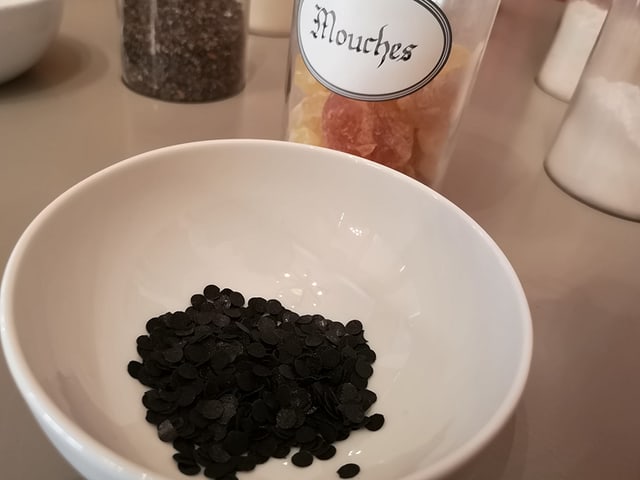 Schälchen mit kleinen schwarzen Punkten. Dahinter ein Glas mit Aufschrift "Mouches". Schälchen mit kleinen schwarzen Punkten. Dahinter ein Glas mit Aufschrift "Mouches".