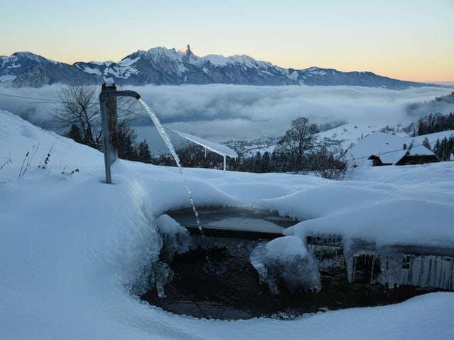 Wasser sprudelt aus einem Hahn. In der Umgebung liegt Schnee, die Dämmerung setzt ein.