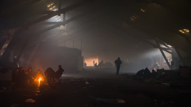 Eine verlassene Industriehalle im Rauch. An mehereren Stellen brennen Feuer, rundum sitzen jeweils ganze Menschengruppen und wärmen sich. Eine verlassene Industriehalle im Rauch. An mehereren Stellen brennen Feuer, rundum sitzen jeweils ganze Menschengruppen und wärmen sich.