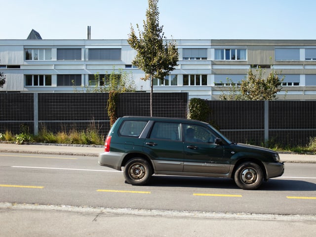Auto fährt auf einer Strasse. Dahinter eine Wand. Im Hintergrund eine Häuserzeile. Auto fährt auf einer Strasse. Dahinter eine Wand. Im Hintergrund eine Häuserzeile.