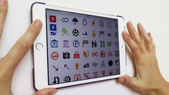 Zwei Hände halten ein Tablet an die Wand. Darauf sind pixelige Emojis zu erkennen.