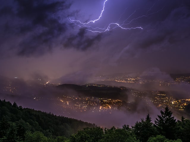 Dunkle Nacht mit vielen Wolken und Blitz am Himmel. Unten dunkler Wald und im Hintergrund ein paar Lichter einer Grossstadt.