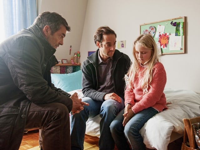 Filmszene: Julia sitzt auf dem Bett in ihrem Zimmer und blick zu Boden. Daneben sitzt ihr Vater, der sie anschaut. Gegenüber, auf einem Stuhl, sitzt Kommissar Flückiger. Filmszene: Julia sitzt auf dem Bett in ihrem Zimmer und blick zu Boden. Daneben sitzt ihr Vater, der sie anschaut. Gegenüber, auf einem Stuhl, sitzt Kommissar Flückiger.