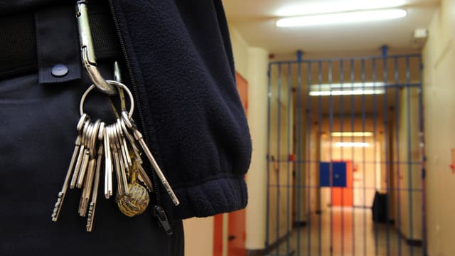 Ein Wärter trägt einen Schlüsselbund. Im Hintergrund ein Gang mit einer Gittertür. Ein Wärter trägt einen Schlüsselbund. Im Hintergrund ein Gang mit einer Gittertür.