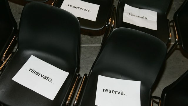 Vier Stühle, die in den vier Landessprachen mit "reserviert" angeschrieben sind. Vier Stühle, die in den vier Landessprachen mit "reserviert" angeschrieben sind.