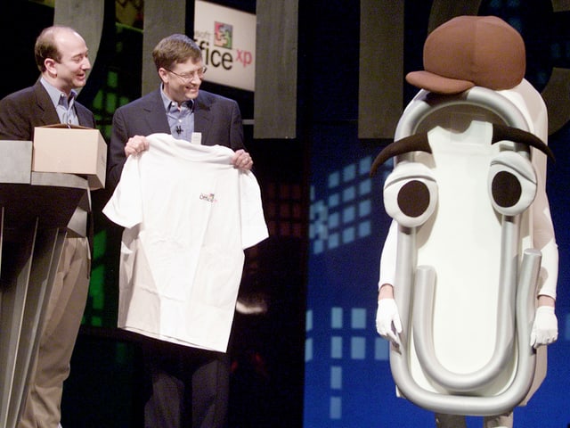 Bill Gates und eine traurige Briefklammer mit Mütze stehen auf einer Bühne, Amazon-CEO Jeff Bezos schaut zu. Bill Gates und eine traurige Briefklammer mit Mütze stehen auf einer Bühne, Amazon-CEO Jeff Bezos schaut zu.