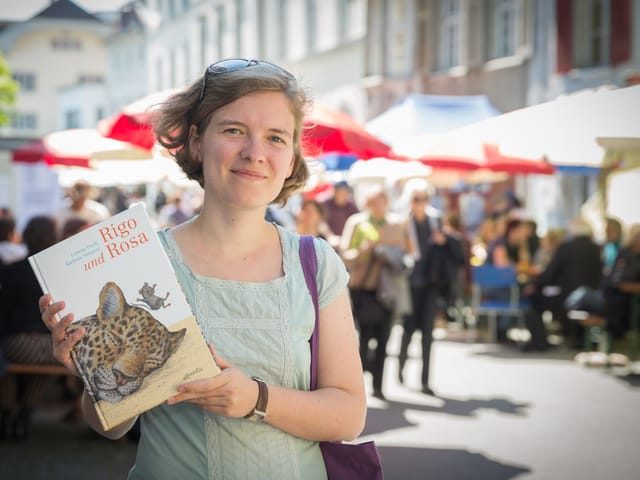 Eine junge Frau steht in einer sonnigen Altstadtgassse mit vielen leuten und hält ein Kinderbuch, auf dem der Kopf eines schlafenden Leoparden zu sehen ist.