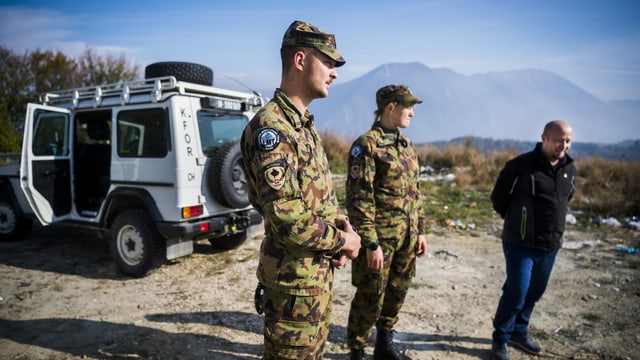 Zwei Swisscoy-Soldaten in Tarntenue stehen neben einem Zivilisten, dahinter ein weisses Fahrzeug, angeschrieben mit KFOR. Zwei Swisscoy-Soldaten in Tarntenue stehen neben einem Zivilisten, dahinter ein weisses Fahrzeug, angeschrieben mit KFOR.