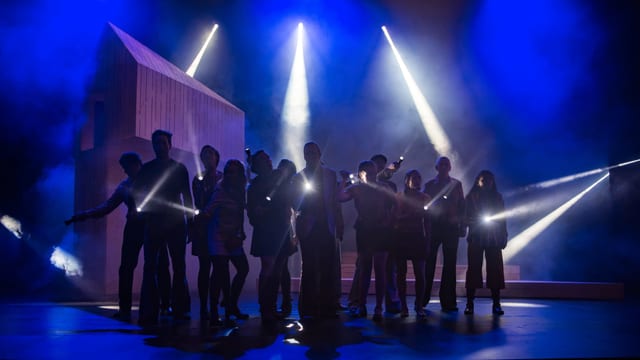 Mehrere Menschen stehen beim Musical Umbra auf der Bühne, man sieht Taschenlampenlicht. Mehrere Menschen stehen beim Musical Umbra auf der Bühne, man sieht Taschenlampenlicht.