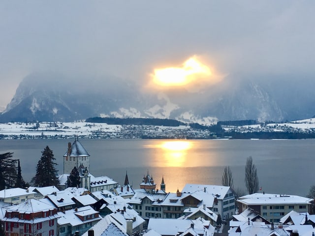 Ein gelber Fleck in der grauen Nebeldecke, auf dem See spiegeln sich die Sonnenstrahlen.