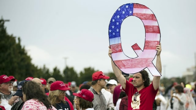 Ina roscha da persunas che portan chapetschas cotschnas pro Trump. In um tegn si in Q zambregià ord cartun. Il Q è en il muster da la bandiera dals Stadis unids da l'America. Ina roscha da persunas che portan chapetschas cotschnas pro Trump. In um tegn si in Q zambregià ord cartun. Il Q è en il muster da la bandiera dals Stadis unids da l'America.