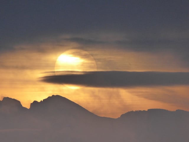 Die Sonne ist als hellgelbe Scheibe hinter dunkelgrauen Wolken zu erkennen. Die Berge zeichnen sich ebenfalls schwarz ab.