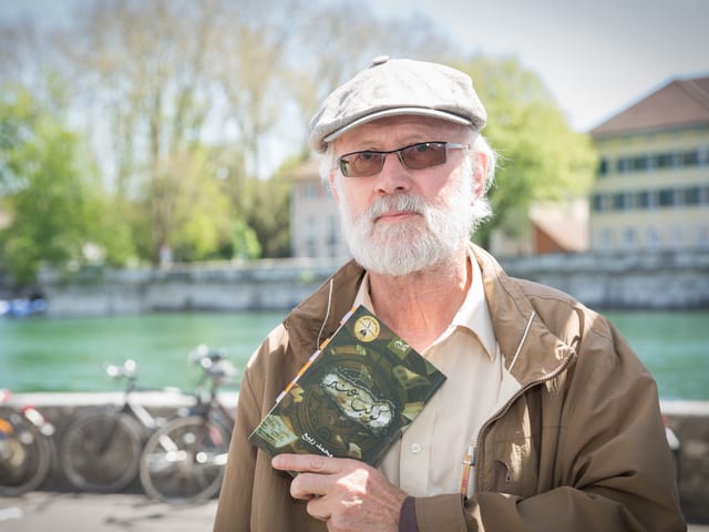 Man mit Bart, Hut und Sonnenbrille steht am Aareufer und hält ein Buch mit arabischen Schriftzeichen.