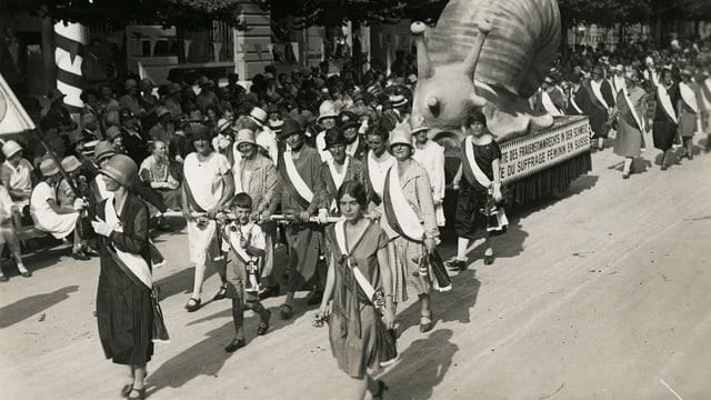 eine schwarz-weiss Fotografie von demonstrierenden Frauen, die eine grosse Schnecke ziehen eine schwarz-weiss Fotografie von demonstrierenden Frauen, die eine grosse Schnecke ziehen