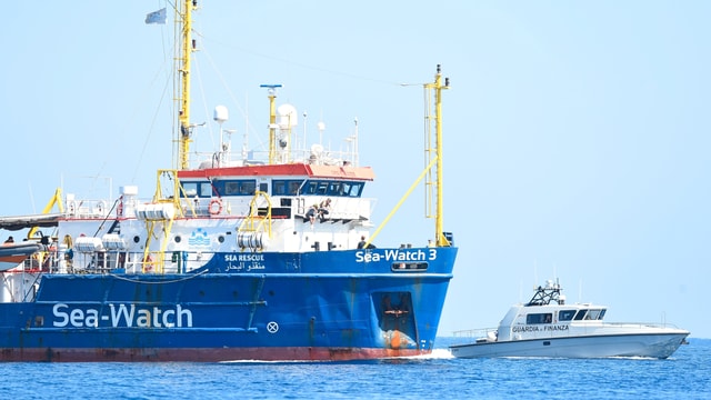 Man sieht ein blaues Rettungsschiff, auf dem "Sea Watch" geschrieben steht und ein kleineres, weisses Boot der Polizei. Man sieht ein blaues Rettungsschiff, auf dem "Sea Watch" geschrieben steht und ein kleineres, weisses Boot der Polizei.