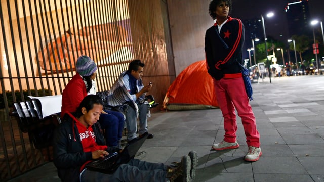 Drei Migranten aus Zentralamerika sitzen am Boden vor einem vergitterten Fenster, einer steht Drei Migranten aus Zentralamerika sitzen am Boden vor einem vergitterten Fenster, einer steht