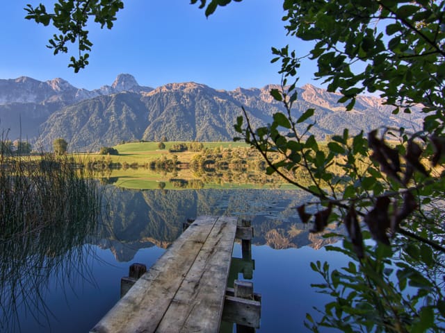 Wolkenloses Wetter an einem kleinen See mit Steg. Die Wasseroberfläche ist glatt und spiegelt Berge, Wälder und grüne Wiesen.