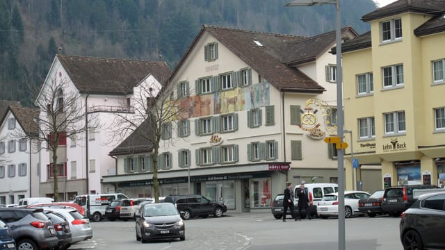 Ein Platz mit parkierten Autos, daneben eine Strasse und ältere Häuser. Ein Platz mit parkierten Autos, daneben eine Strasse und ältere Häuser.