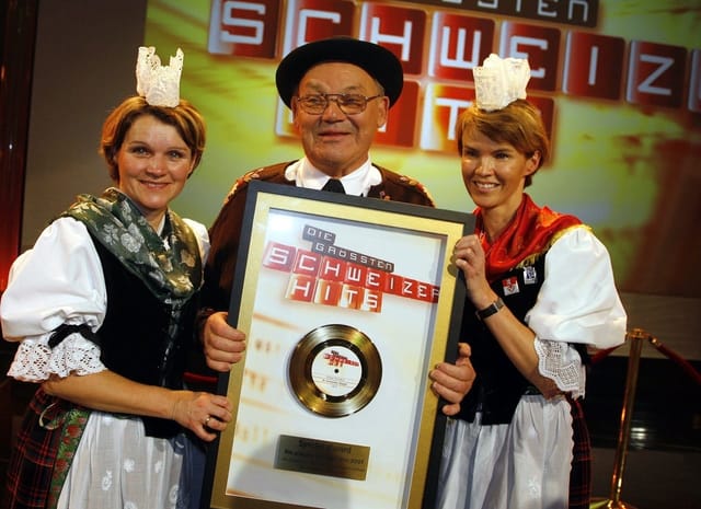 Mann hält goldene Schallplatte in die Kamera. Zwei Frauen in Trachten stehen ihm zur Seite. Mann hält goldene Schallplatte in die Kamera. Zwei Frauen in Trachten stehen ihm zur Seite.