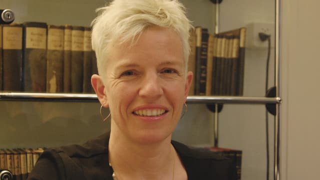 Eine Frau mit blondem, kurzem Haar steht vor einem Glasregal mit alten Büchern. Eine Frau mit blondem, kurzem Haar steht vor einem Glasregal mit alten Büchern.