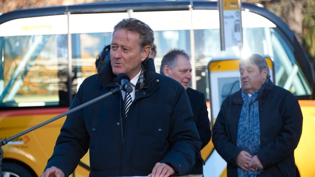 Ein Mann an einem Rednerpult, dahinter ein Postauto Ein Mann an einem Rednerpult, dahinter ein Postauto