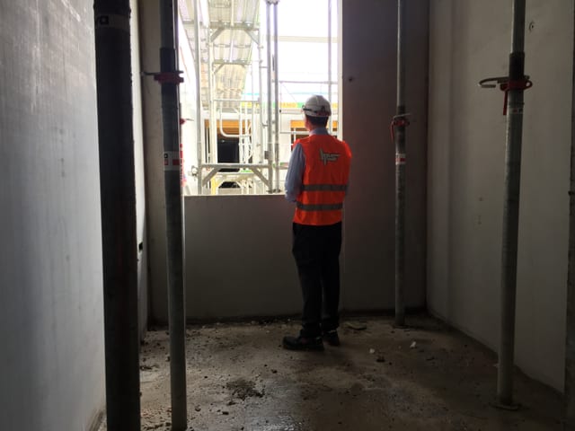 Mann in oranger Leuchtweste steht in engem Raum an einer Fensterlucke Mann in oranger Leuchtweste steht in engem Raum an einer Fensterlucke