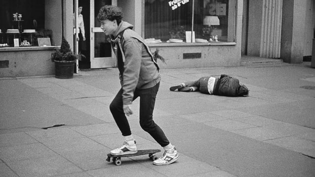 Ein junger Skater fährt an einem Mann vorbei, der auf der Strasse liegt.