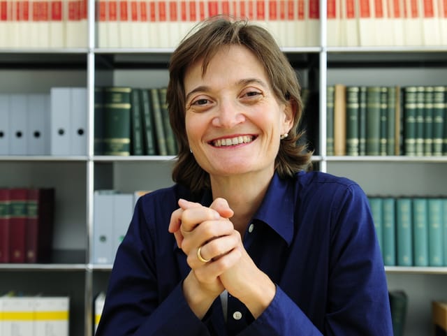 Frau mit schulterlangen braunen Haaren und blauer Bluse sitzt vor einem Bücherregal. Frau mit schulterlangen braunen Haaren und blauer Bluse sitzt vor einem Bücherregal.